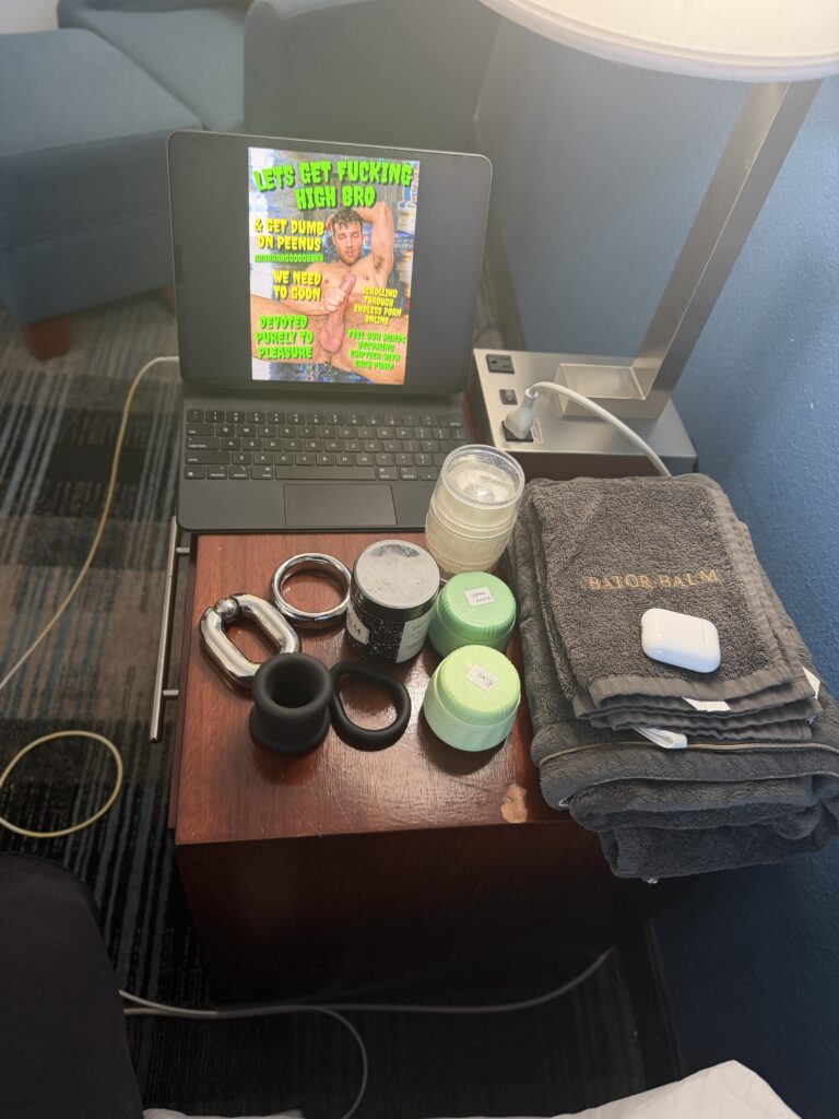 Hotel side table covered with lube, cockrings and ball weights, rags, and an iPad with a batecap on it